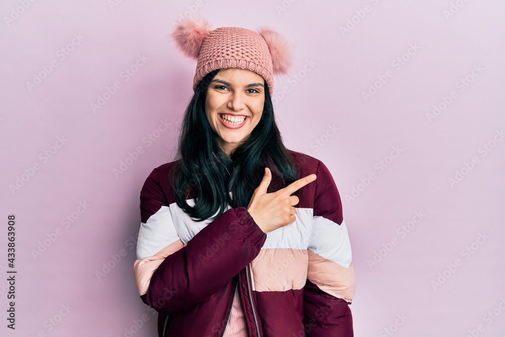 Young hispanic woman wearing wool winter sweater and cap cheerful with a smile of face pointing with hand and finger up to the side with happy and natural expression on face Young hispanic woman wearing wool winter sweater and cap cheerful with a smile of face pointing with hand and finger up to the side with happy and natural expression on face