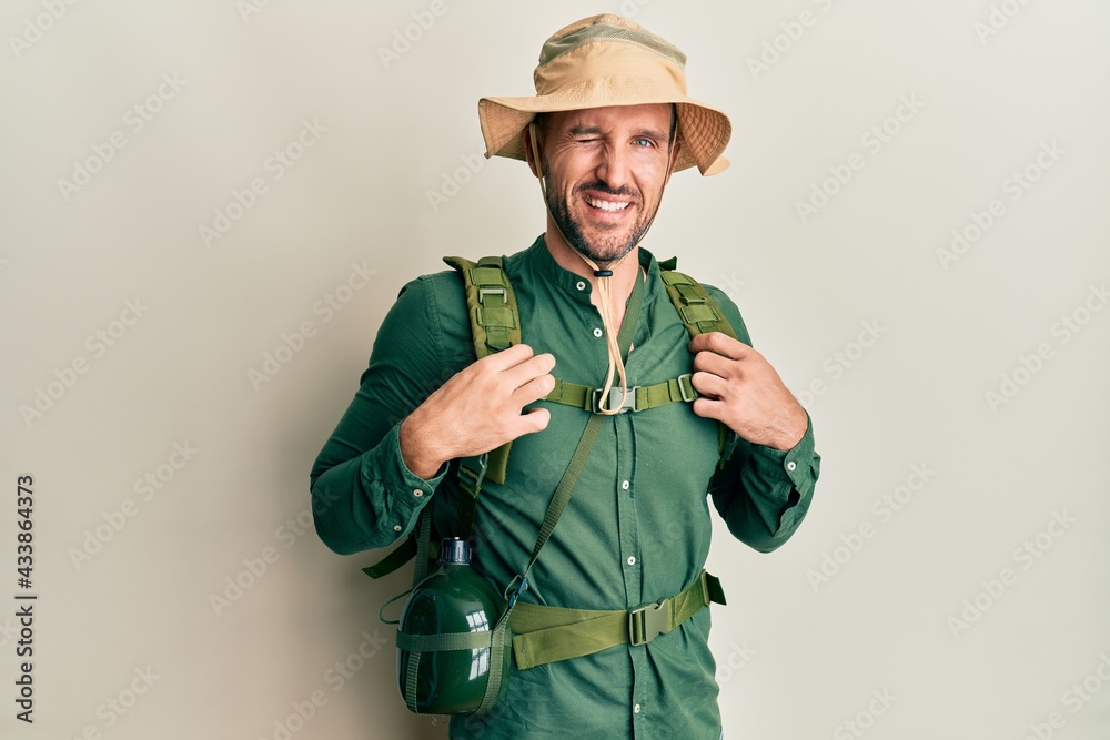 Handsome man with beard wearing explorer hat and backpack winking looking at the camera with sexy expression, cheerful and happy face. Handsome man with beard wearing explorer hat and backpack winking looking at the camera with sexy expression, cheerful and happy face.