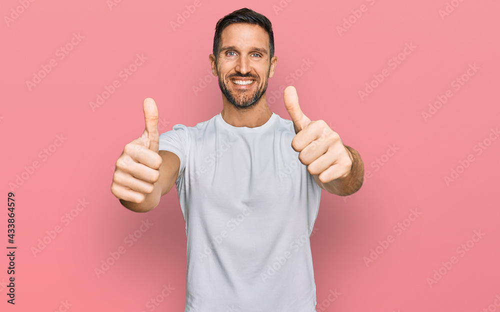 Handsome man with beard wearing casual white t shirt approving doing positive gesture with hand, thumbs up smiling and happy for success. winner gesture. Handsome man with beard wearing casual white t shirt approving doing positive gesture with hand, thumbs up smiling and happy for success. winner gesture.