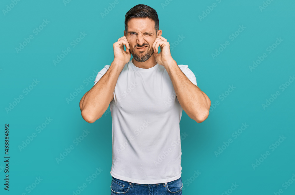Handsome man with beard wearing casual white t shirt covering ears with fingers with annoyed expression for the noise of loud music. deaf concept. Handsome man with beard wearing casual white t shirt covering ears with fingers with annoyed expression for the noise of loud music. deaf concept.