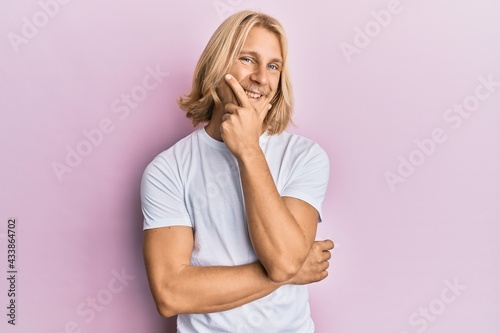 Caucasian young man with long hair wearing casual white t shirt looking confident at the camera smiling with crossed arms and hand raised on chin. thinking positive.