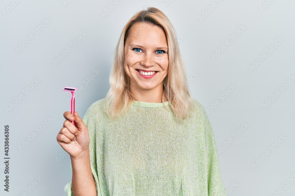 Beautiful caucasian blonde woman holding razor looking positive and happy standing and smiling with a confident smile showing teeth