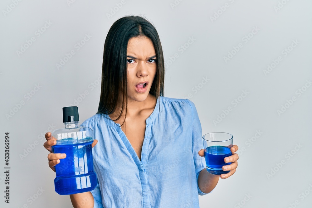 Young hispanic girl holding mouthwash for fresh breath in shock face, looking skeptical and sarcastic, surprised with open mouth Young hispanic girl holding mouthwash for fresh breath in shock face, looking skeptical and sarcastic, surprised with open mouth