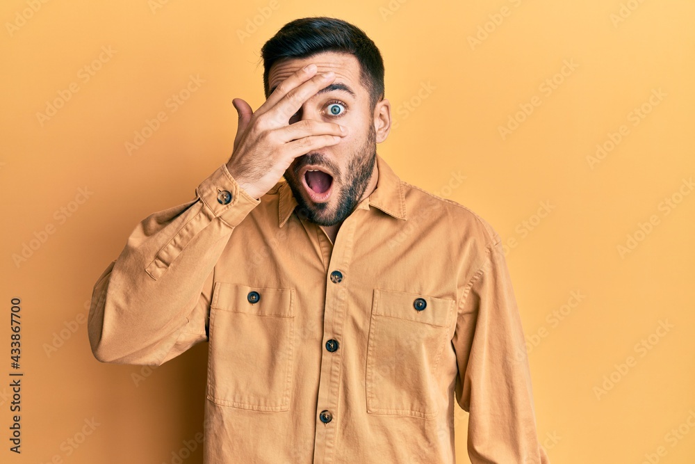 Young hispanic man wearing casual clothes peeking in shock covering face and eyes with hand, looking through fingers afraid Young hispanic man wearing casual clothes peeking in shock covering face and eyes with hand, looking through fingers afraid