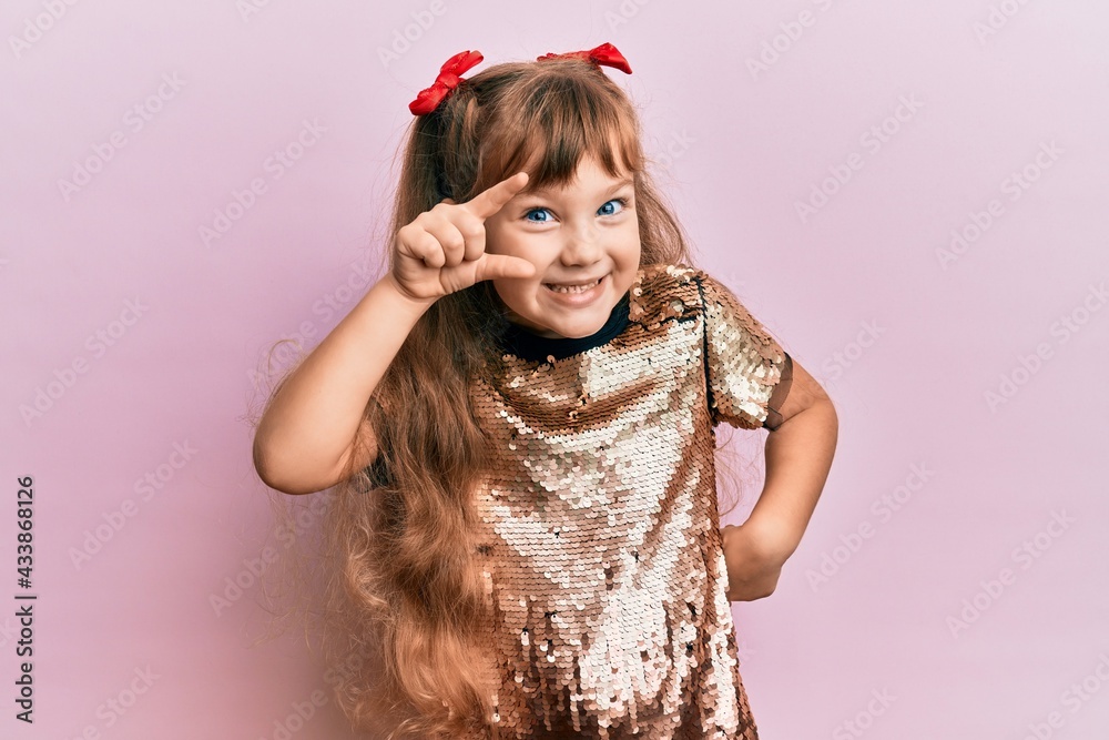 Little caucasian girl kid wearing festive sequins dress smiling and confident gesturing with hand doing small size sign with fingers looking and the camera. measure concept. Little caucasian girl kid wearing festive sequins dress smiling and confident gesturing with hand doing small size sign with fingers looking and the camera. measure concept.