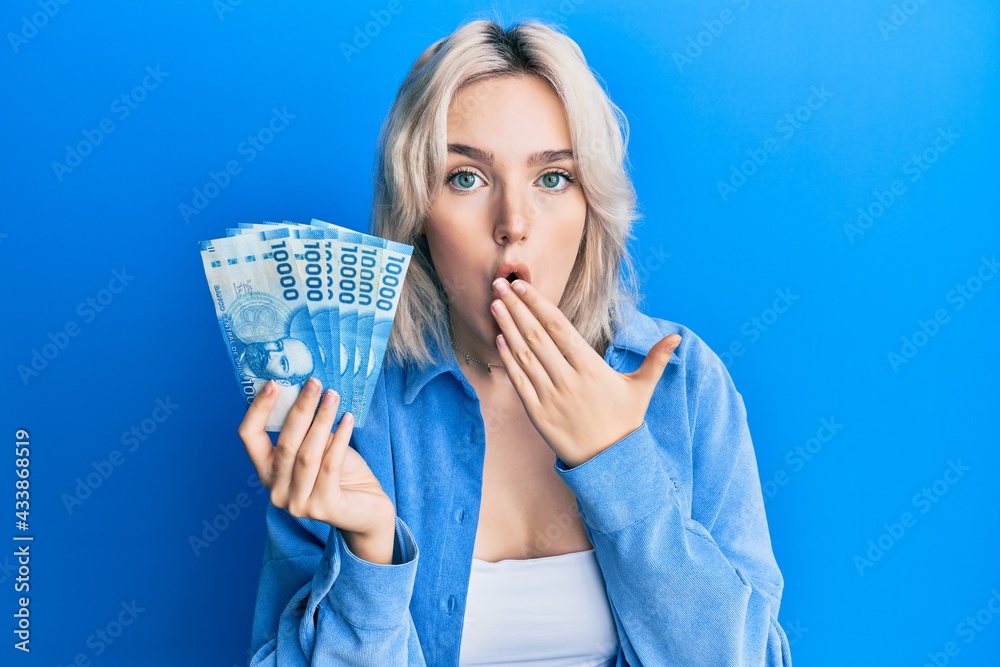 Young blonde girl holding 1000 chilean pesos covering mouth with hand, shocked and afraid for mistake. surprised expression