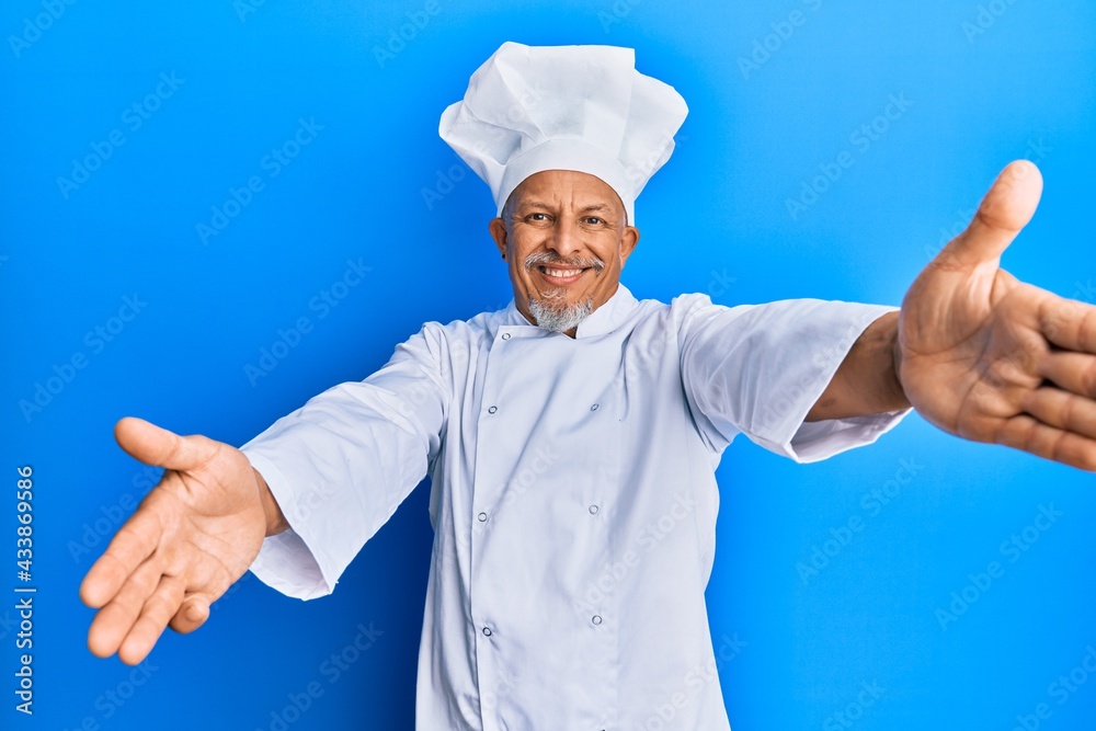Middle age grey-haired man wearing professional cook uniform and hat looking at the camera smiling with open arms for hug. cheerful expression embracing happiness. Middle age grey-haired man wearing professional cook uniform and hat looking at the camera smiling with open arms for hug. cheerful expression embracing happiness.