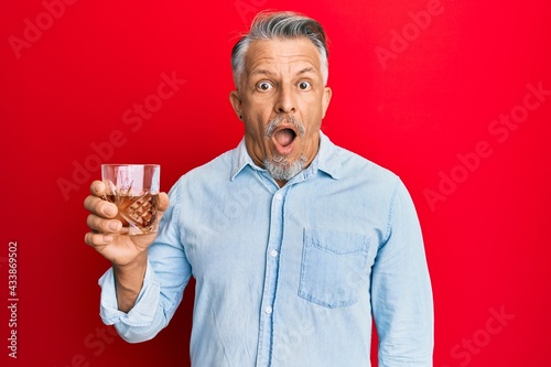Middle age grey-haired man drinking glass of whisky scared and amazed with open mouth for surprise, disbelief face