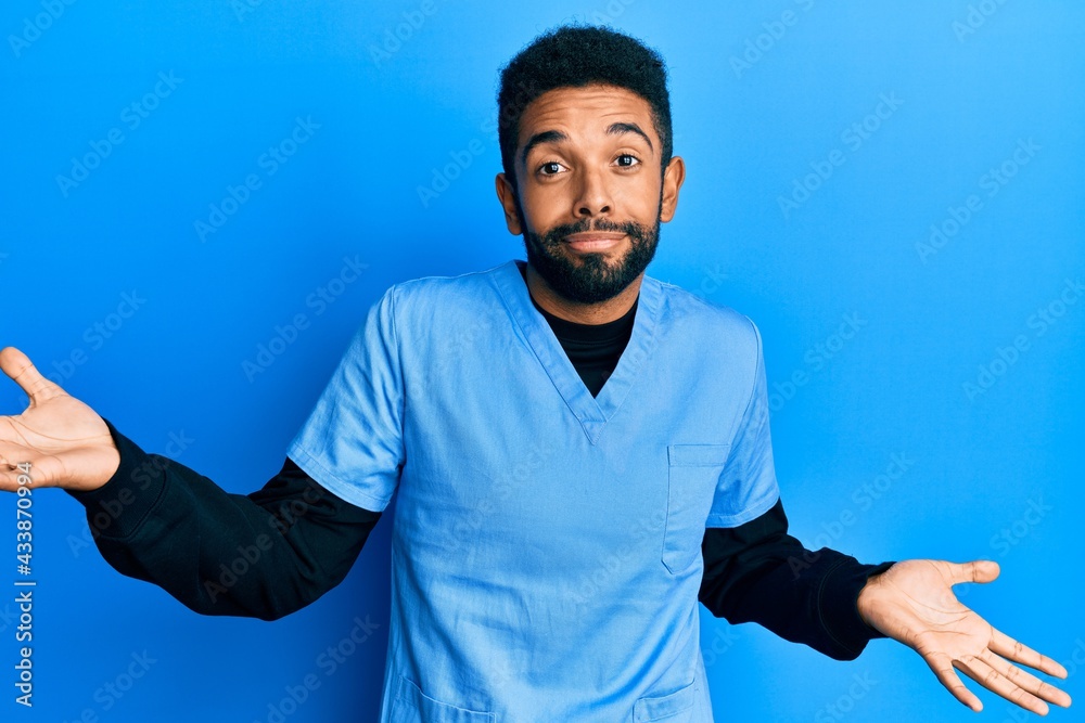 Handsome hispanic man with beard wearing blue male nurse uniform clueless and confused expression with arms and hands raised. doubt concept.