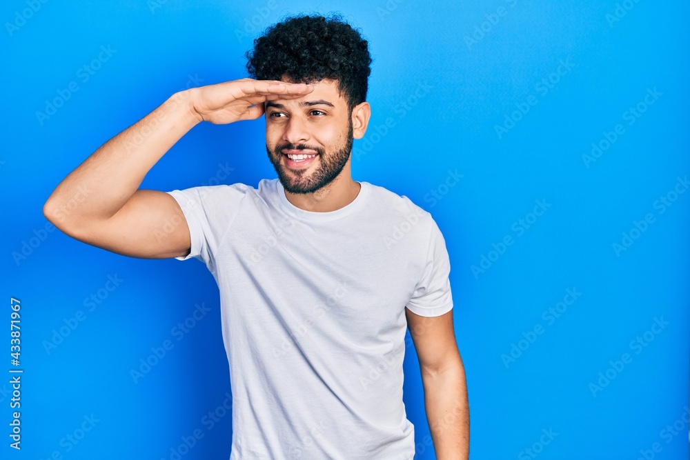 Young arab man with beard wearing casual white t shirt very happy and smiling looking far away with hand over head. searching concept. Young arab man with beard wearing casual white t shirt very happy and smiling looking far away with hand over head. searching concept.