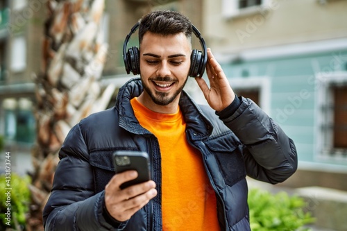 Handsome hispanic man with beard smiling happy and confident at the city wearing winter coat listening to music from smartphone