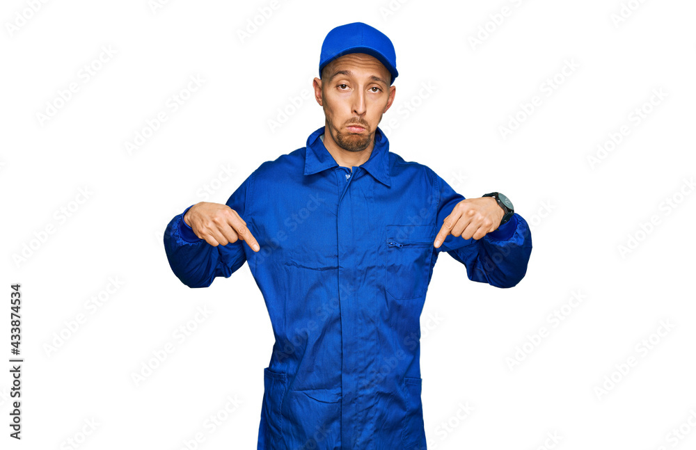 Bald man with beard wearing builder jumpsuit uniform pointing down looking sad and upset, indicating direction with fingers, unhappy and depressed.