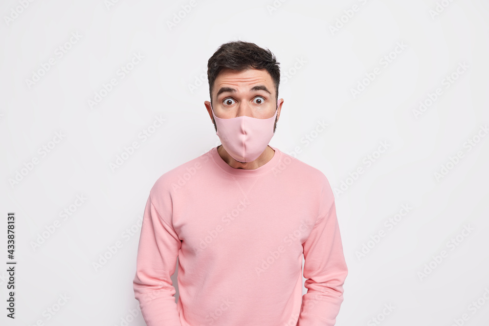 Personal protection and quarantine concept. Startled young man wears face mask to protect from coronavirus hears shocking news dressed in jumper isolated over white background. Viral infection Personal protection and quarantine concept. Startled young man wears face mask to protect from coronavirus hears shocking news dressed in jumper isolated over white background. Viral infection