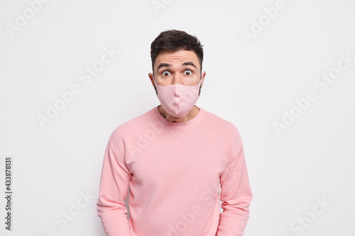 Personal protection and quarantine concept. Startled young man wears face mask to protect from coronavirus hears shocking news dressed in jumper isolated over white background. Viral infection