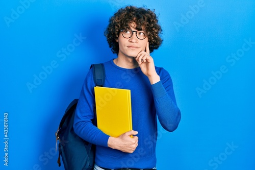 Handsome young man wearing student backpack and holding book serious face thinking about question with hand on chin, thoughtful about confusing idea