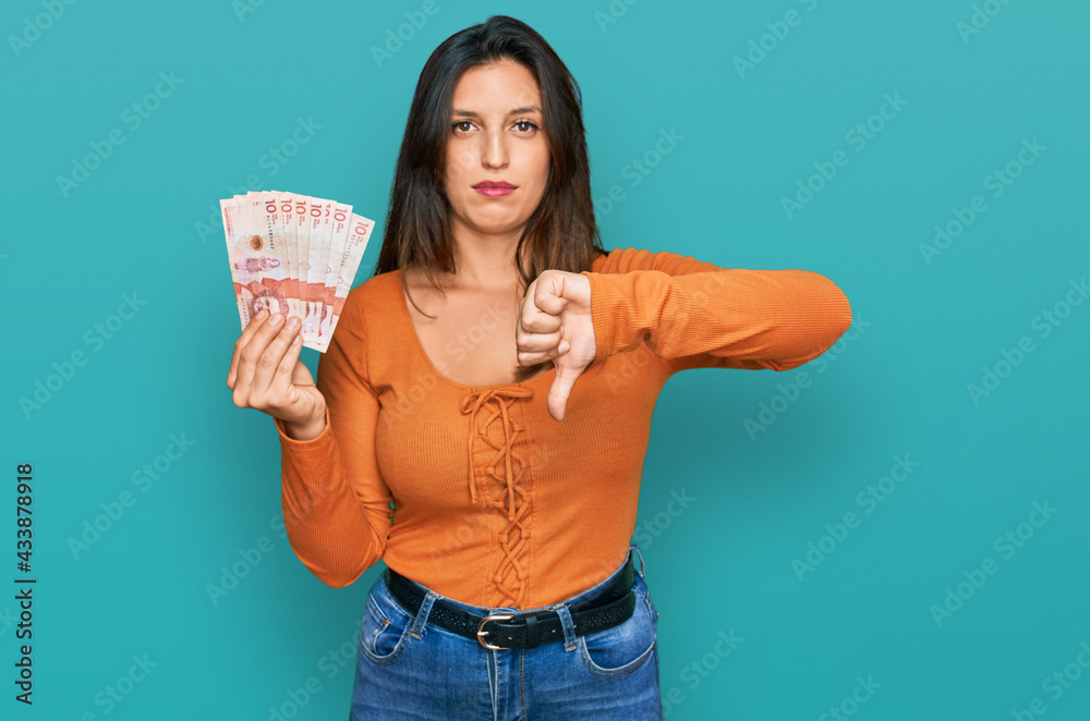 Beautiful hispanic woman holding 10 colombian pesos banknotes with angry face, negative sign showing dislike with thumbs down, rejection concept Beautiful hispanic woman holding 10 colombian pesos banknotes with angry face, negative sign showing dislike with thumbs down, rejection concept