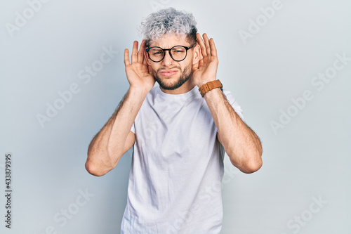 Young hispanic man with modern dyed hair wearing white t shirt and glasses trying to hear both hands on ear gesture, curious for gossip. hearing problem, deaf