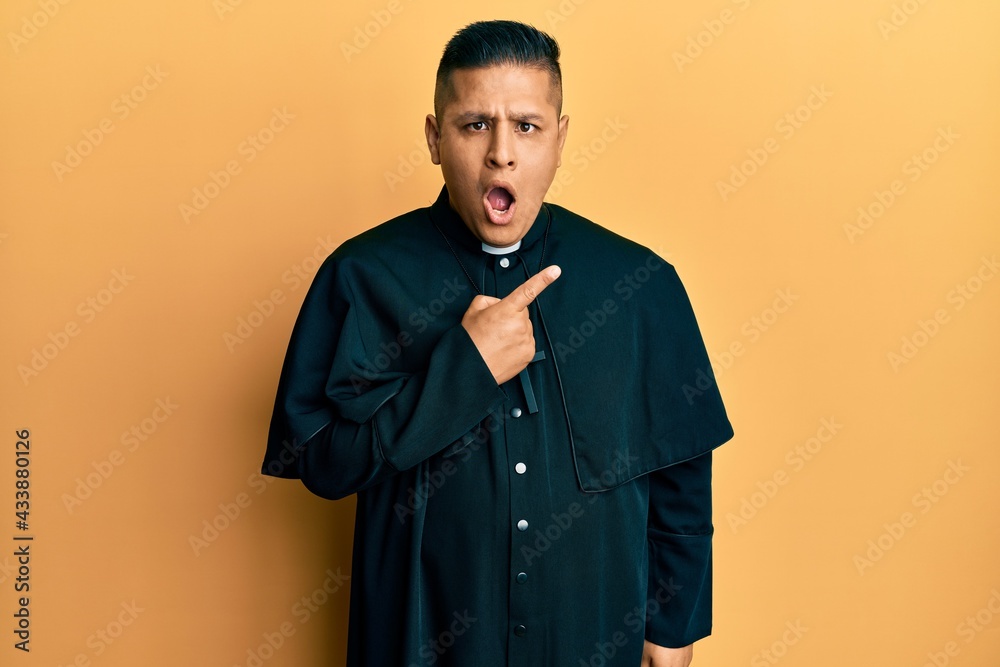 Young latin priest man standing over yellow background surprised pointing with finger to the side, open mouth amazed expression. - obrazy, fototapety, plakaty 