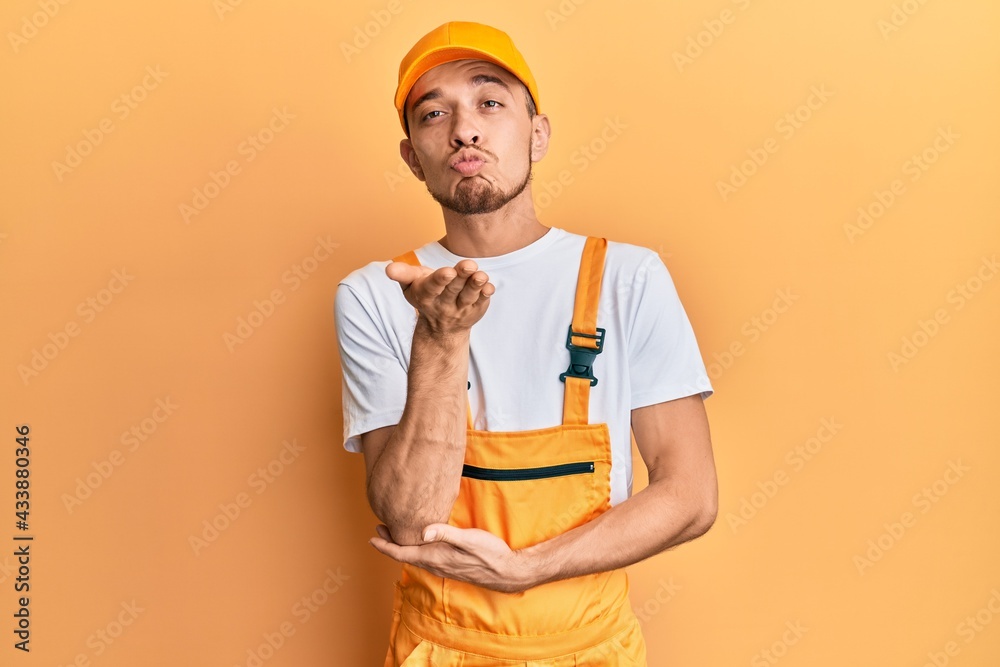 Hispanic young man wearing handyman uniform looking at the camera blowing a kiss with hand on air being lovely and sexy. love expression. Hispanic young man wearing handyman uniform looking at the camera blowing a kiss with hand on air being lovely and sexy. love expression.