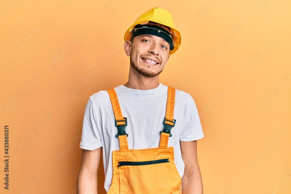 Hispanic young man wearing builder uniform and safety hardhat with a happy and cool smile on face. lucky person.