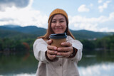 Portrait image of a young woman holding and giving coffee cup while traveling mountains and the lake