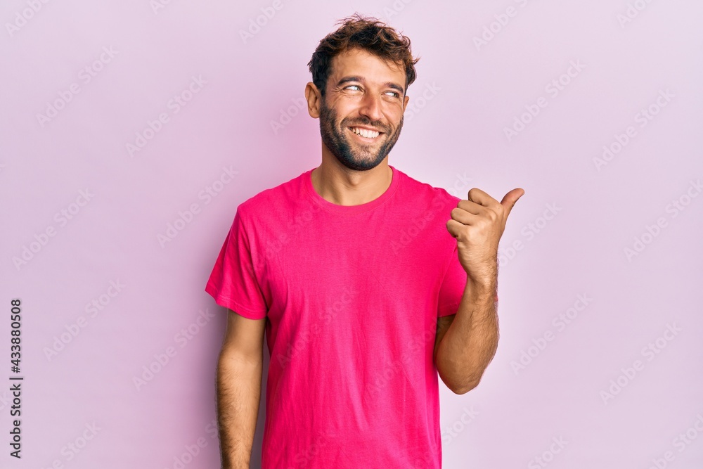Handsome man with beard wearing casual pink tshirt over pink background smiling with happy face looking and pointing to the side with thumb up. Handsome man with beard wearing casual pink tshirt over pink background smiling with happy face looking and pointing to the side with thumb up.