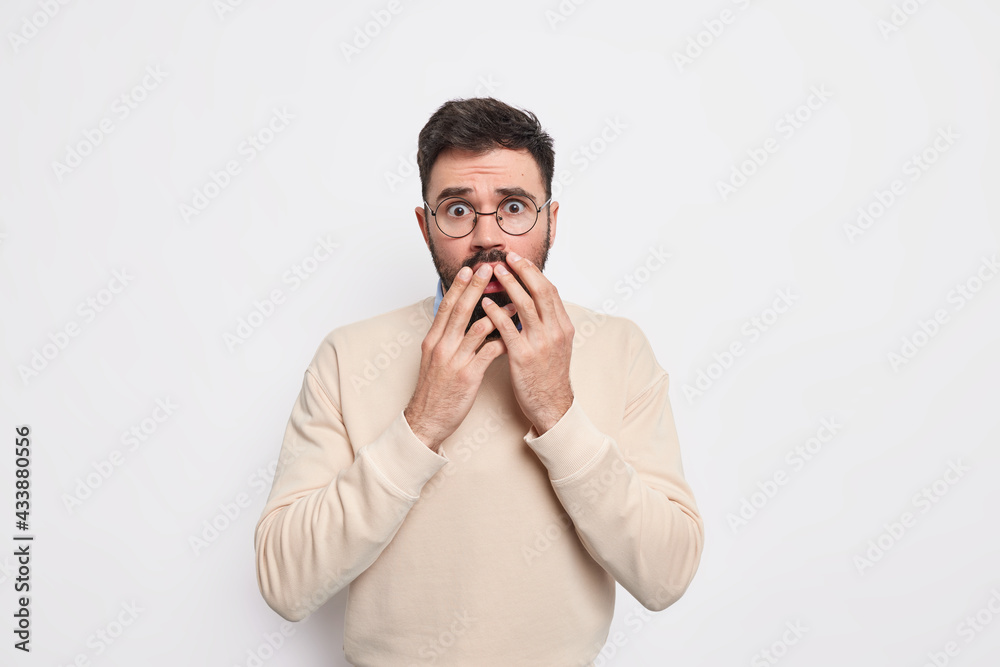 Speechless scared man looks with fright at camera trembles from fear keeps hands on mouth wears round spectacles and jumper isolated over white background. Anxious worried male model feels insecure Speechless scared man looks with fright at camera trembles from fear keeps hands on mouth wears round spectacles and jumper isolated over white background. Anxious worried male model feels insecure