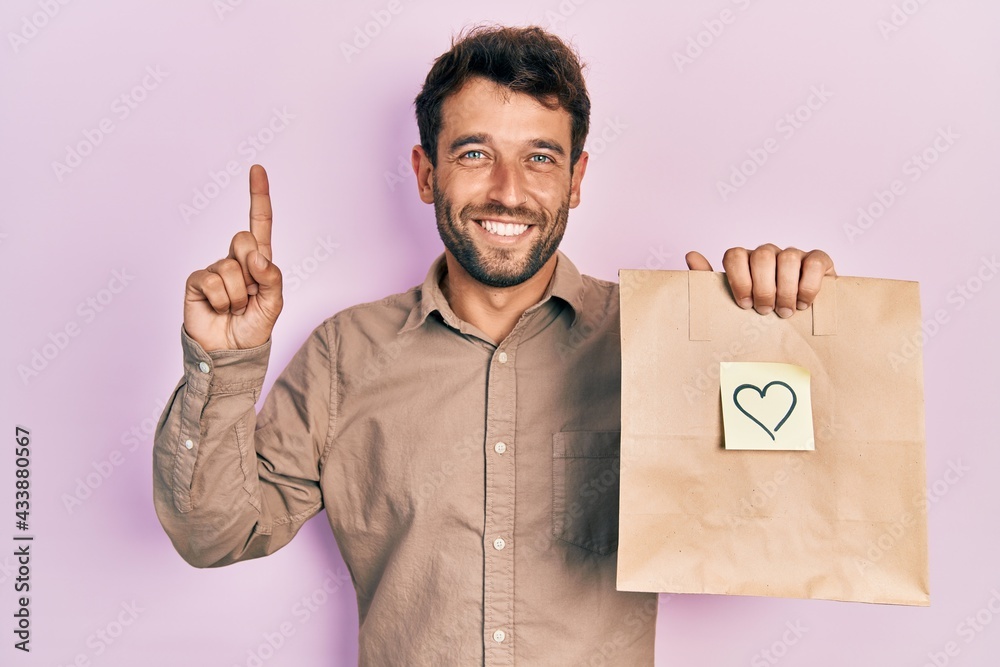 Handsome man with beard holding delivery paper bag with heart reminder smiling amazed and surprised and pointing up with fingers and raised arms. Handsome man with beard holding delivery paper bag with heart reminder smiling amazed and surprised and pointing up with fingers and raised arms.