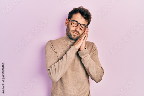 Handsome man with beard wearing turtleneck sweater and glasses sleeping tired dreaming and posing with hands together while smiling with closed eyes. © Krakenimages.com