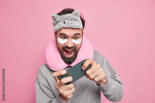 Cheerful bearded man plays video games on smartphone before going to bed wears sleepmask and comfortable pillow around neck beauty patches addicted to modern technologies tries to pass hard level. © Wayhome Studio