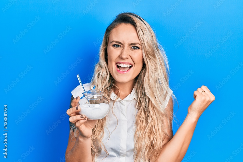 Beautiful young blonde woman holding bowl with sugar screaming proud, celebrating victory and success very excited with raised arms Beautiful young blonde woman holding bowl with sugar screaming proud, celebrating victory and success very excited with raised arms