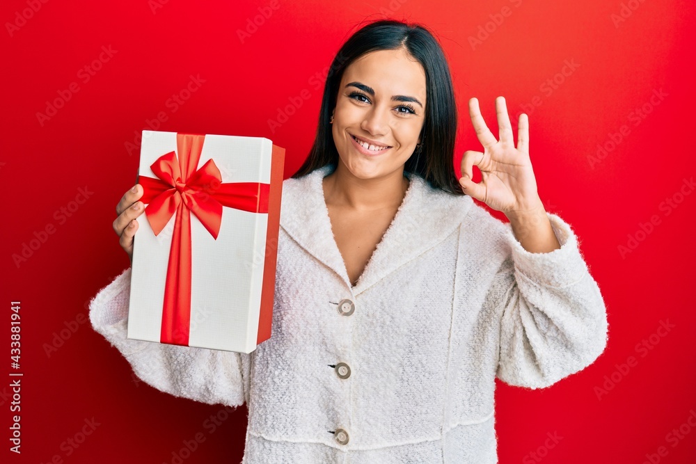 Young brunette woman holding gift doing ok sign with fingers, smiling friendly gesturing excellent symbol Young brunette woman holding gift doing ok sign with fingers, smiling friendly gesturing excellent symbol