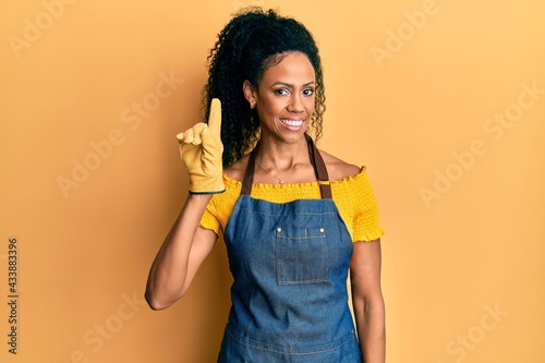 Middle age african american woman wearing professional apron pointing finger up with successful idea. exited and happy. number one.