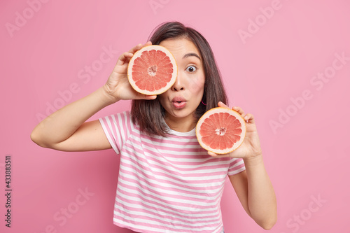 Surprised young Asian woman poses with citrus fruit indoor holds two halves of fresh grapefruit keeps to healthy nutrition has shocked expression dressed in t shirt isolated over pink background