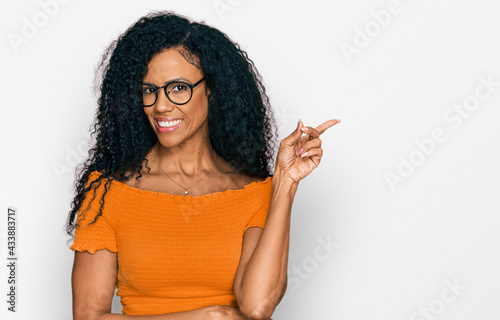 Middle age african american woman wearing casual clothes and glasses with a big smile on face, pointing with hand and finger to the side looking at the camera.