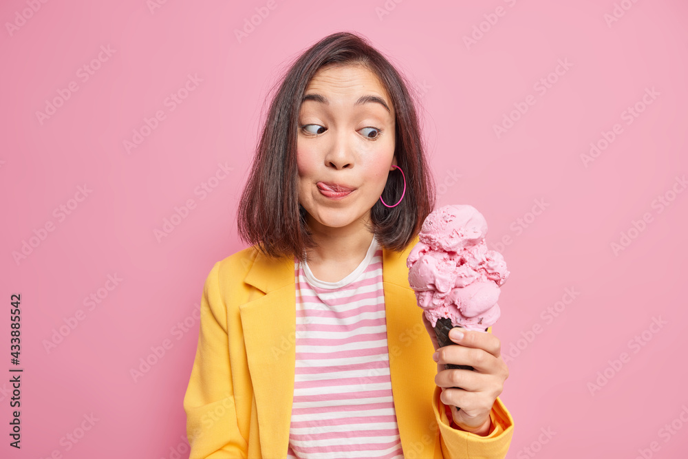 Pretty young dark haired Asian woman looks at appetizing yummy ice cream licks lips with tongue cannot wait until eating delicious summer dessert dressed in fashionable clothes. Sweet tooth concept Pretty young dark haired Asian woman looks at appetizing yummy ice cream licks lips with tongue cannot wait until eating delicious summer dessert dressed in fashionable clothes. Sweet tooth concept