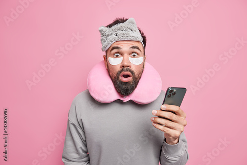 Amazed bearded man reacts on astonishing news looks with disbelief stands speechless holds breath uses smartphone surfs internet prepares for having nap wears sleepmask neckpillow isolated on pink