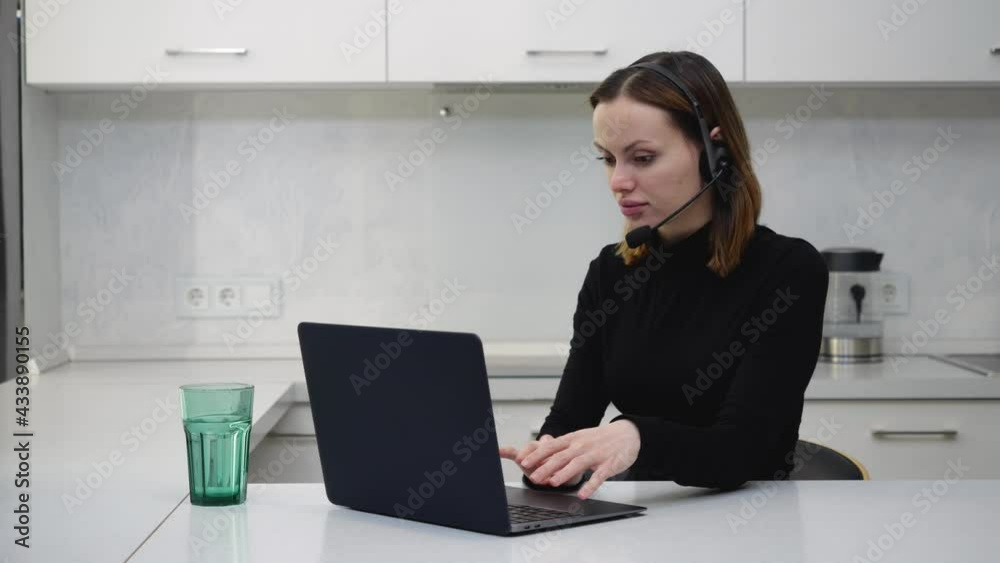Young girl in black sweater and headset headphones talking to a customer while working behind a laptop. High quality 4k footage Young girl in black sweater and headset headphones talking to a customer while working behind a laptop. High quality 4k footage