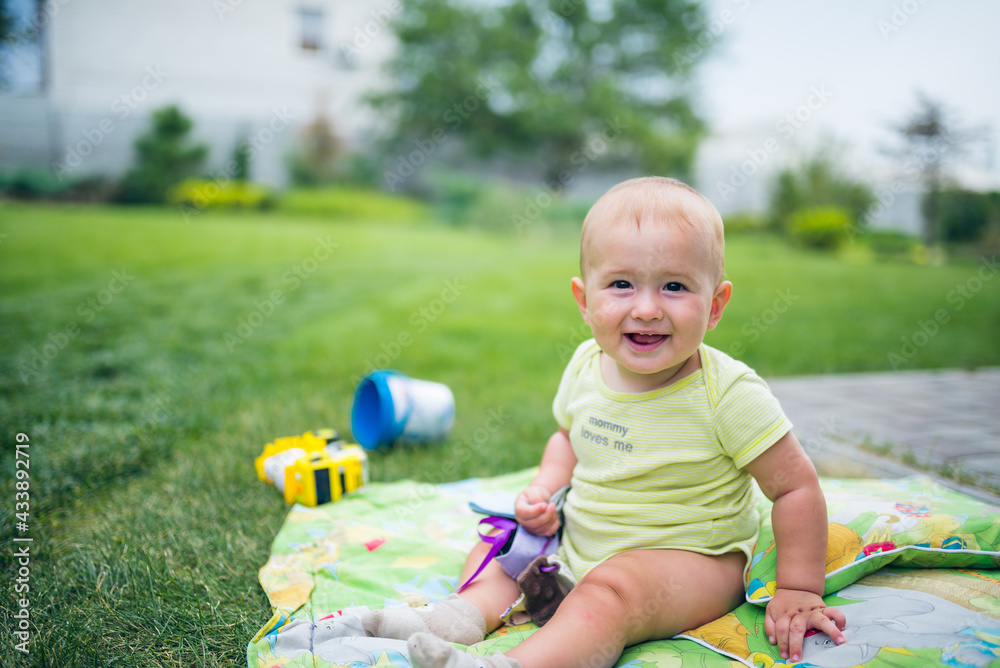 Fototapeta premium A babyboy is sitting on a green blanket in the garden and smiling. Two first teeth show up. A huge space of green grass is in the background.
