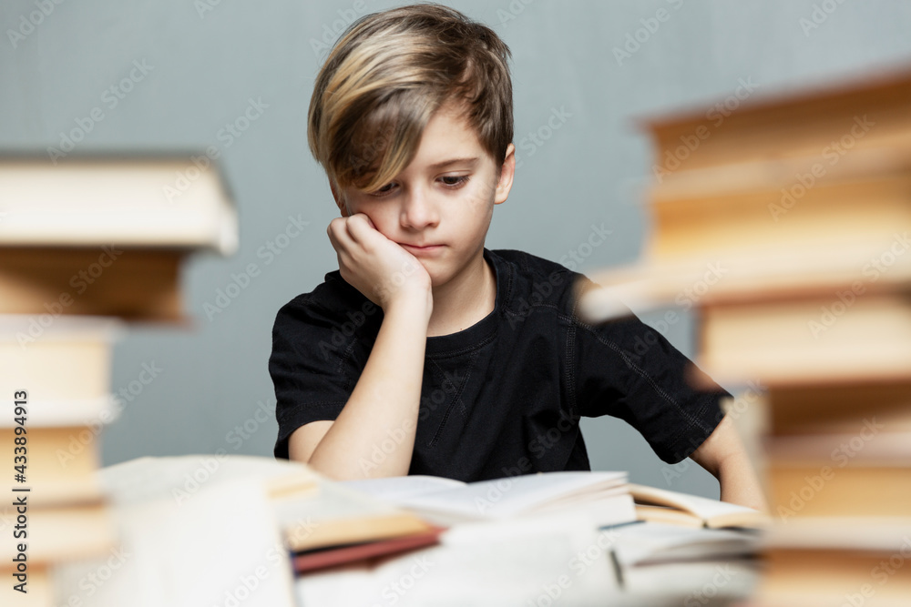 A pensive boy of 9-10 years old sits at a table with books. Gray background. Exams and learning difficulties.