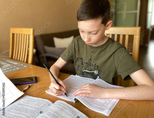 Schoolboy in a t-shirt with a textbook at the desk. Teenager studies, does his homework. Boy with a book and pencil