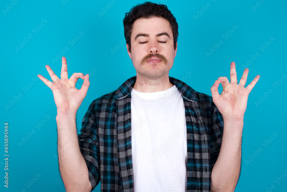 Naklejka premium young Caucasian man with moustache wearing plaid shirt against blue wall doing yoga, keeping eyes closed, holding fingers in mudra gesture. Meditation, religion and spiritual practices.