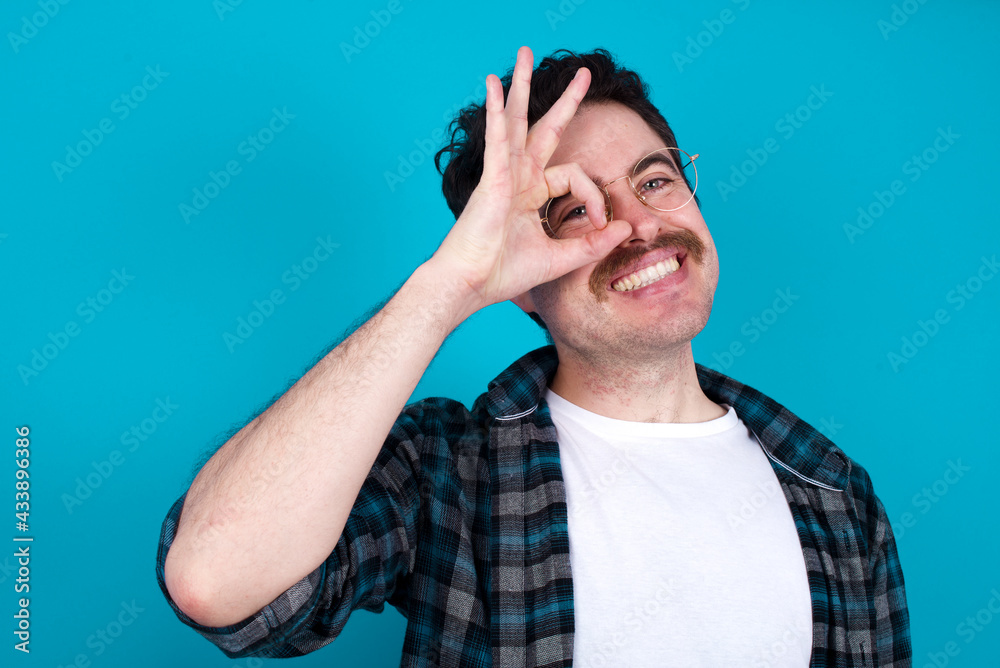 young Caucasian man with moustache wearing plaid shirt against blue wall with happy face smiling doing ok sign with hand on eye looking through finger. young Caucasian man with moustache wearing plaid shirt against blue wall with happy face smiling doing ok sign with hand on eye looking through finger.