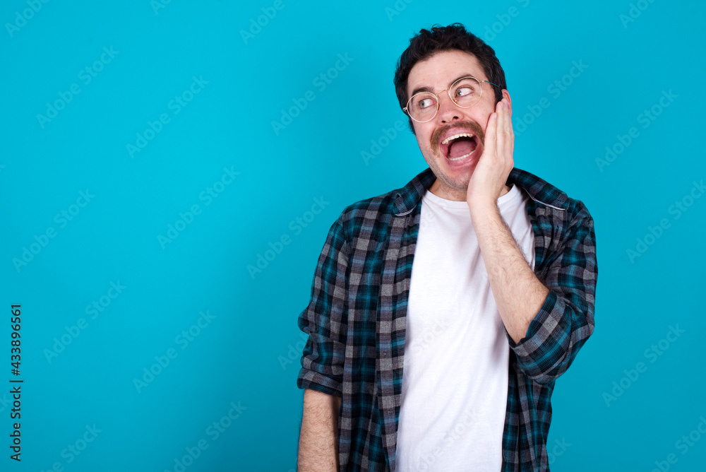 young Caucasian man with moustache wearing plaid shirt against blue wall excited looking to the side hand on face. Advertisement and amazement concept.