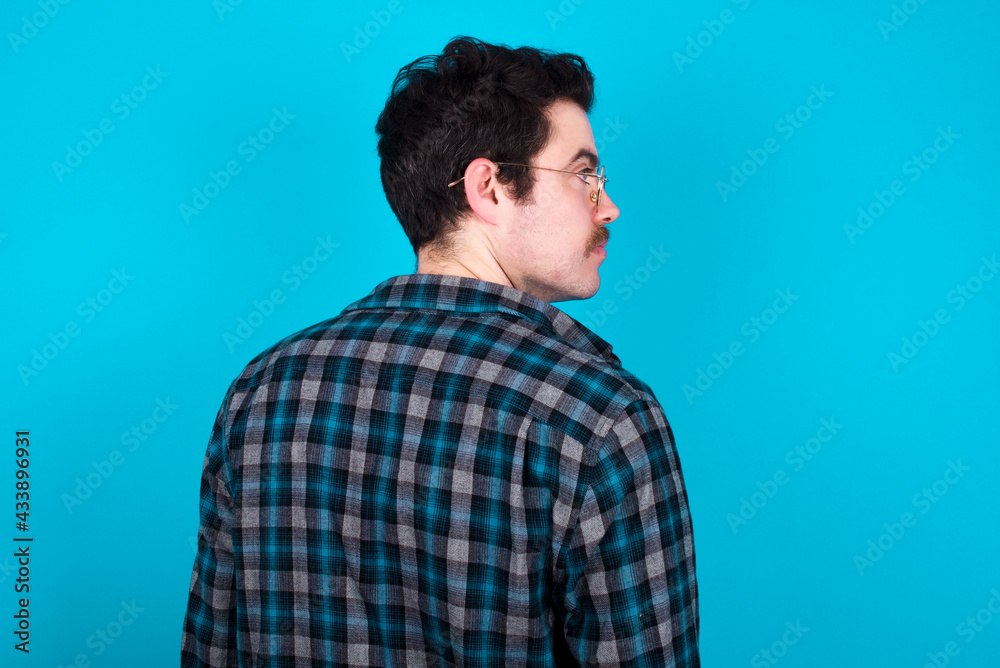 The back view of young Caucasian man with moustache wearing plaid shirt against blue wall Studio Shoot. The back view of young Caucasian man with moustache wearing plaid shirt against blue wall Studio Shoot.