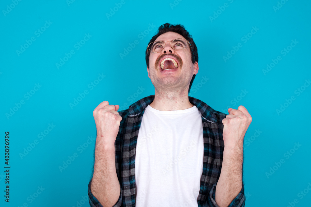 young Caucasian man with moustache wearing plaid shirt against blue wall looks with excitement up, keeps hands raised, notices something unexpected.