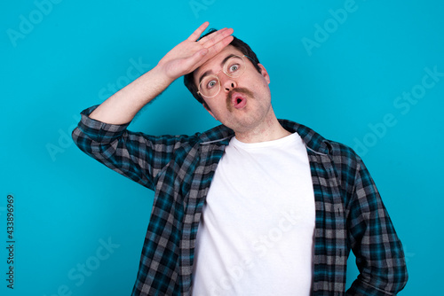 Foto young Caucasian man with moustache wearing plaid shirt against blue, wiping forehead with hand making phew gesture, expressing relief feels happy that he prevented huge disaster