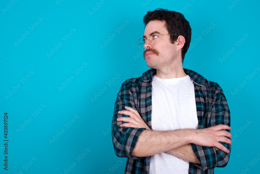 Image of upset young Caucasian man with moustache wearing plaid shirt against blue wall with arms crossed. Looking with disappointed expression aside after listening to bad news. Image of upset young Caucasian man with moustache wearing plaid shirt against blue wall with arms crossed. Looking with disappointed expression aside after listening to bad news.