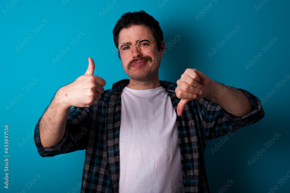 young Caucasian man with moustache wearing plaid shirt against blue wall feeling unsure making good bad sign. Displeased and unimpressed. young Caucasian man with moustache wearing plaid shirt against blue wall feeling unsure making good bad sign. Displeased and unimpressed.