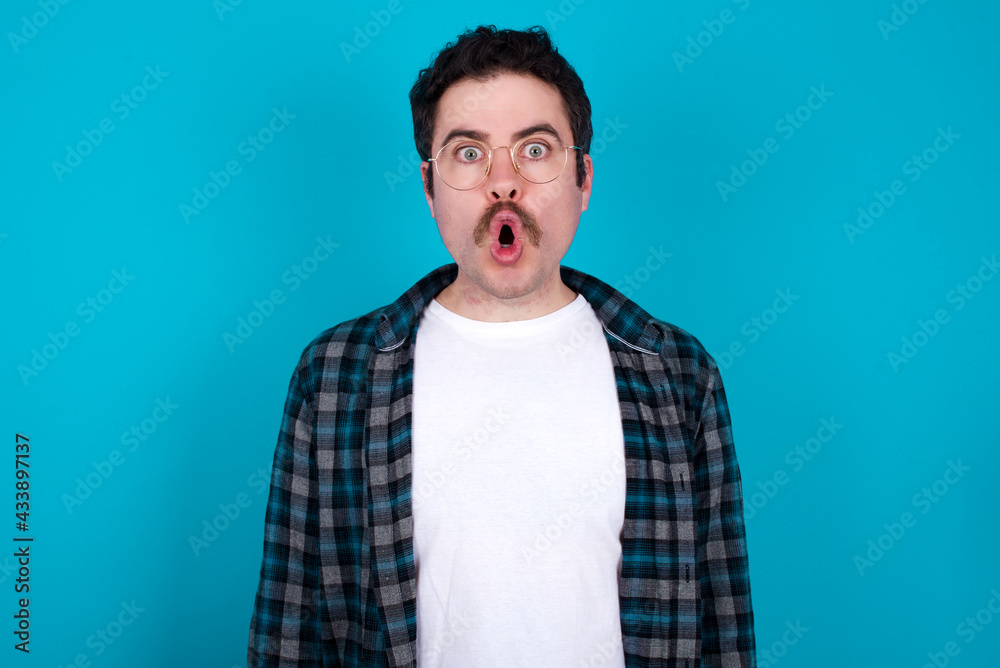 Emotional attractive young Caucasian man with moustache wearing plaid shirt agains with opened mouth expresses great surprise and fright, stares at camera. Unexpected shocking news and human reaction. Emotional attractive young Caucasian man with moustache wearing plaid shirt agains with opened mouth expresses great surprise and fright, stares at camera. Unexpected shocking news and human reaction.